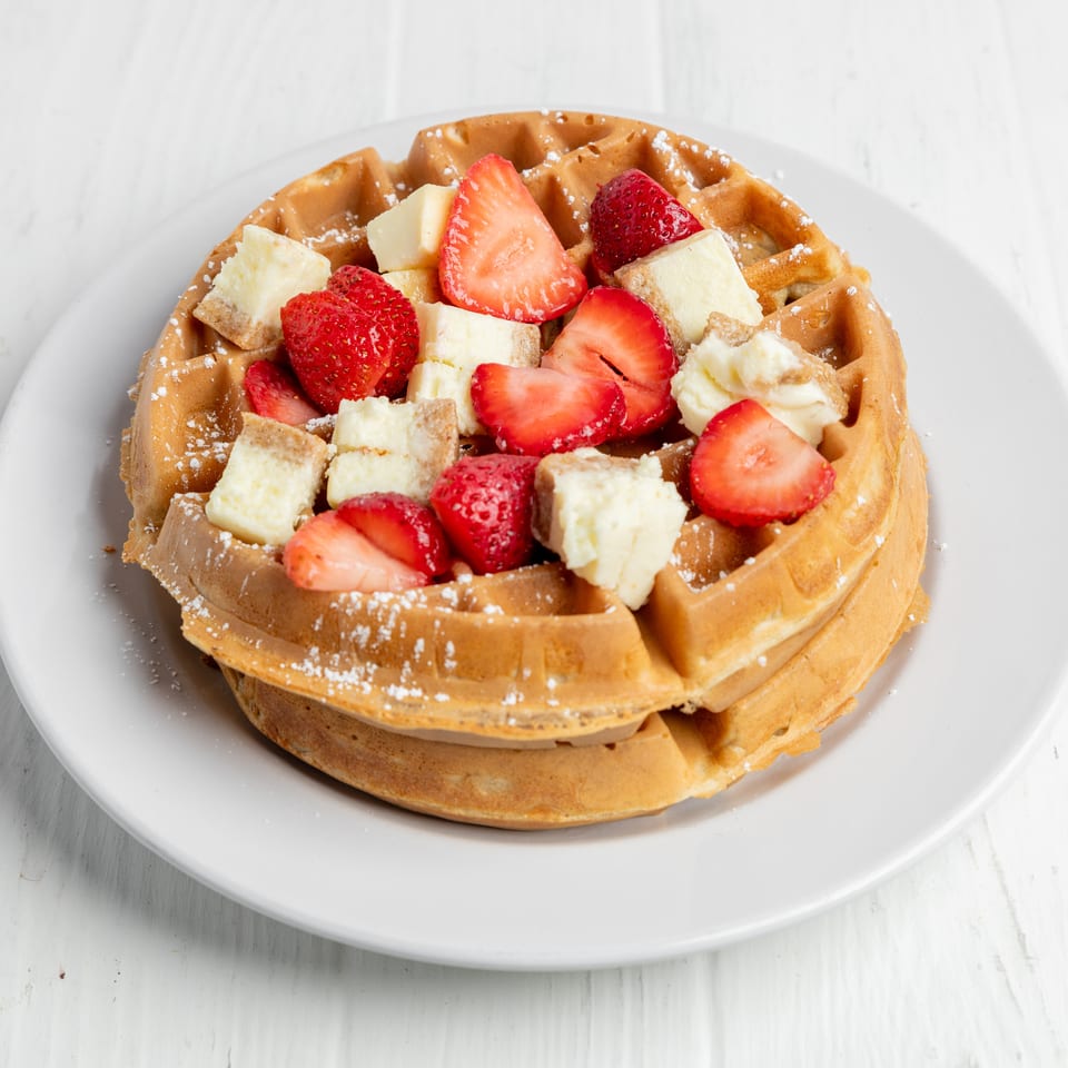 Golden waffle topped with sliced strawberries, banana pieces, and powdered sugar on a white plate.