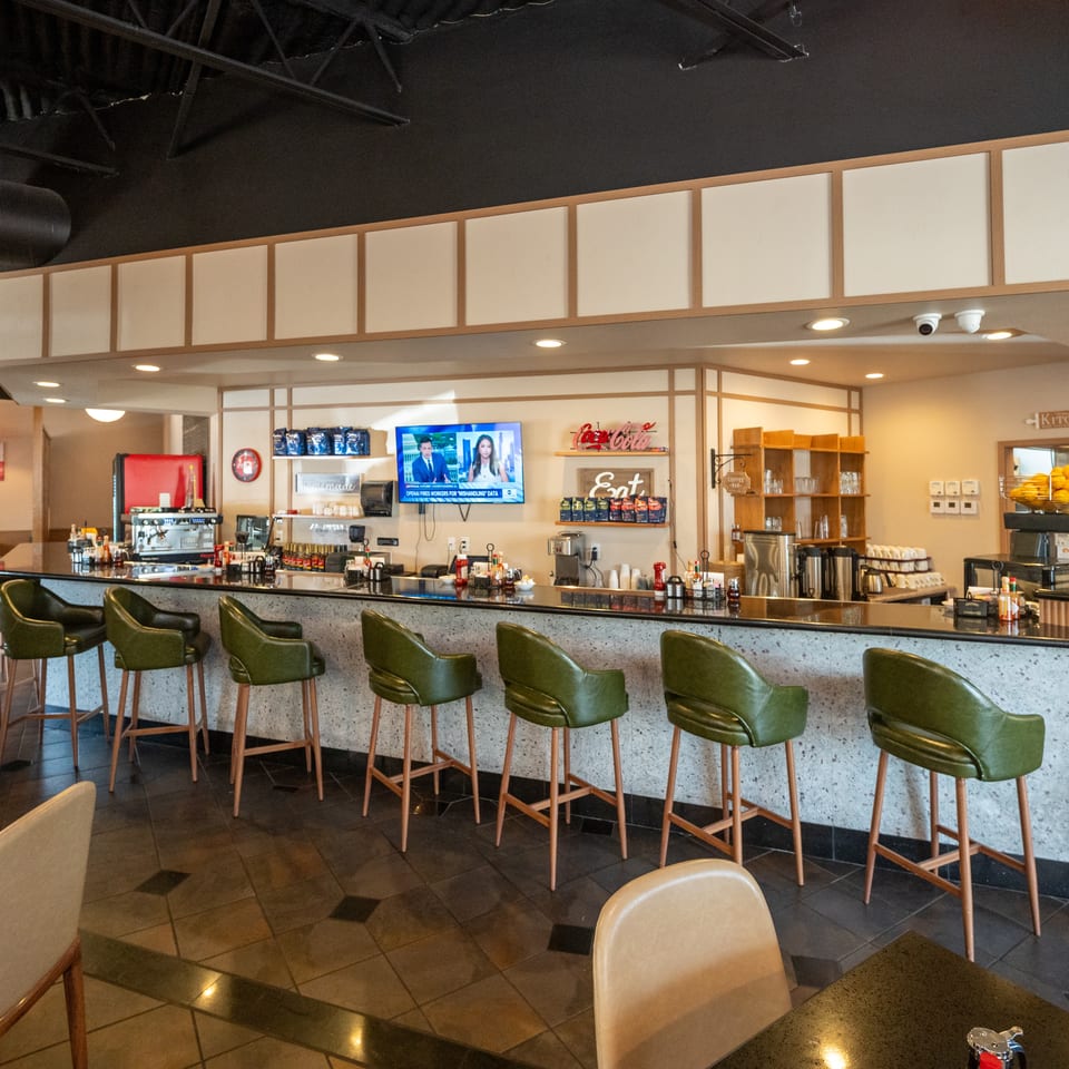 Modern restaurant interior with a long counter, green bar stools, beverage station, television, and warm contemporary decor.