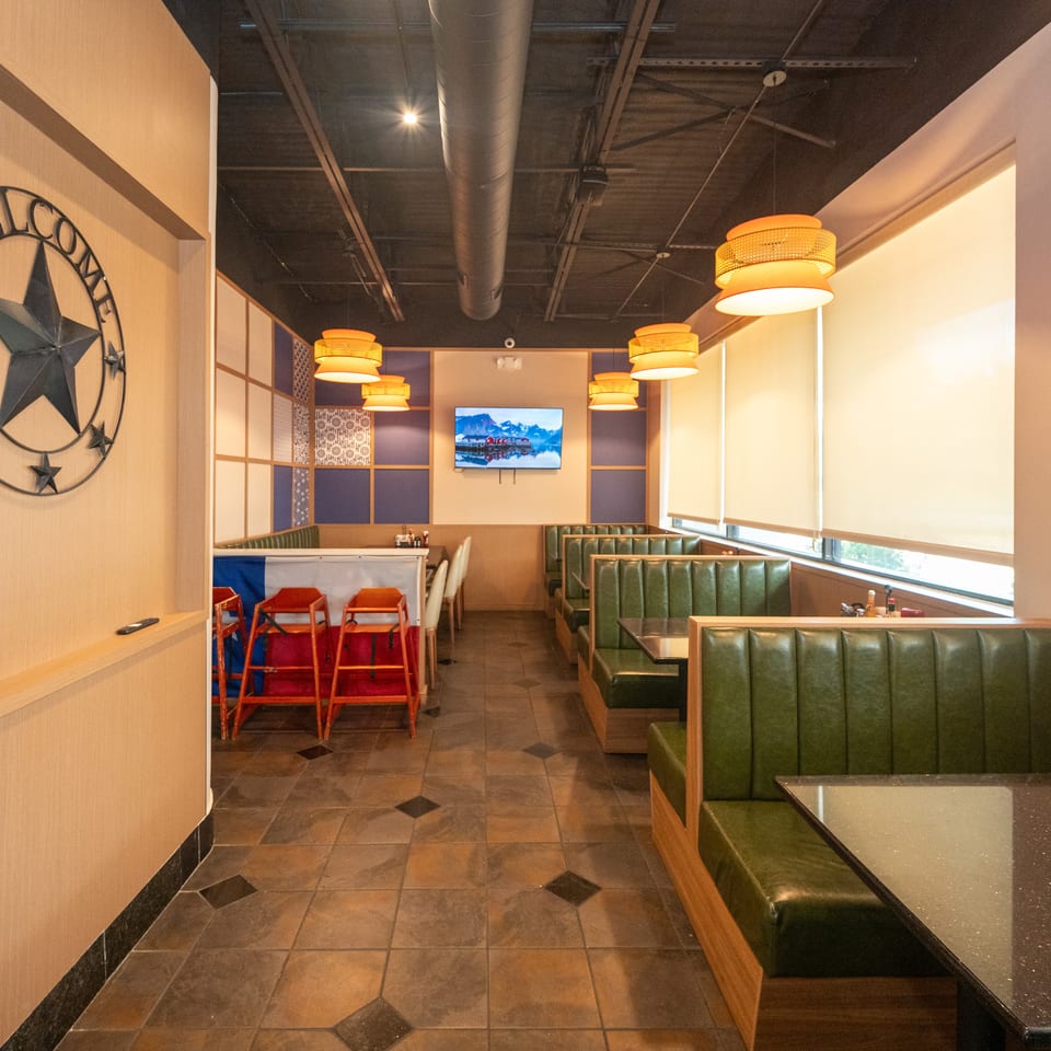 Warmly lit restaurant dining area with green leather booths, pendant lights, tiled floors, wall art, and a mounted television.