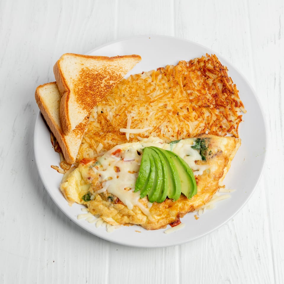 Cheesy omelet topped with sliced avocado, served with crispy hash browns and toasted bread on a white plate.