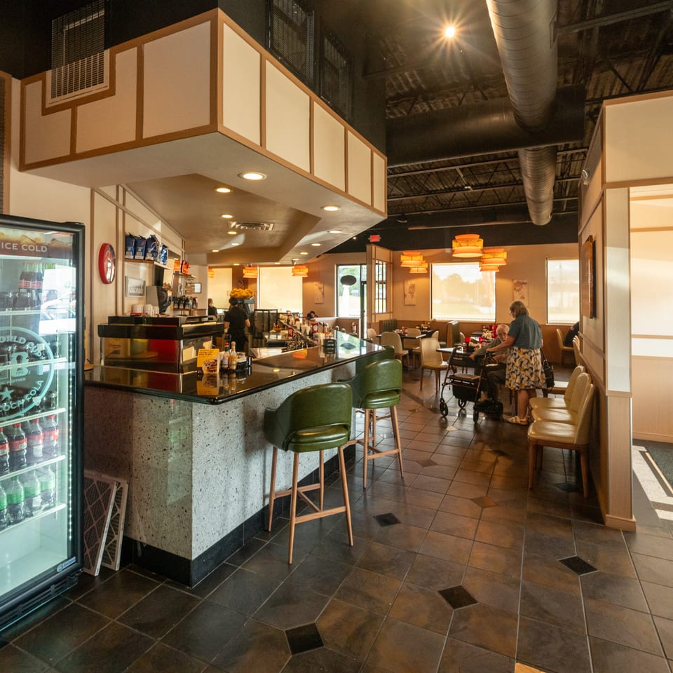 Warmly lit restaurant interior with a counter, green barstools, booths, pendant lights, beverage cooler, and diners.