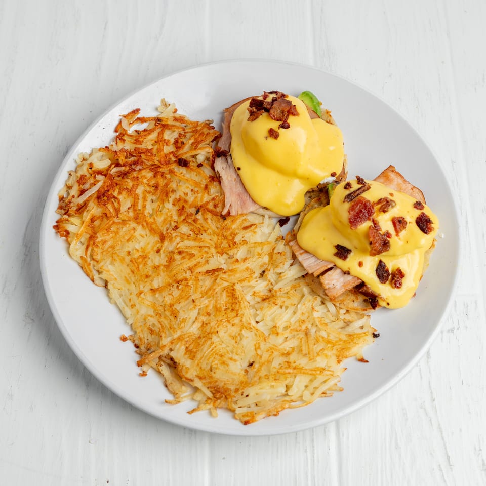 Plate of eggs Benedict with ham, avocado, bacon, and hollandaise sauce served alongside crispy hash browns.