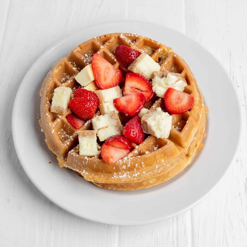 Golden waffle topped with sliced strawberries, banana pieces, and powdered sugar on a white plate.