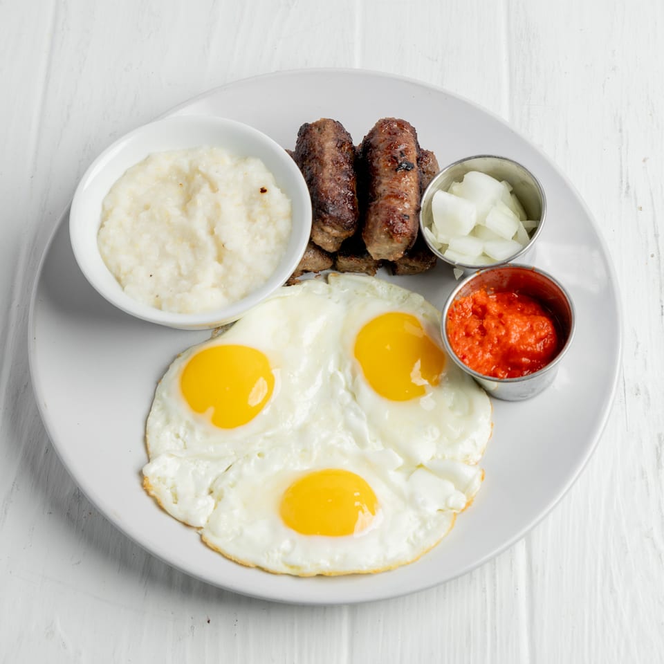Plate of fried eggs, grilled sausage, creamy grits, chopped onions, and red sauce on a white table.