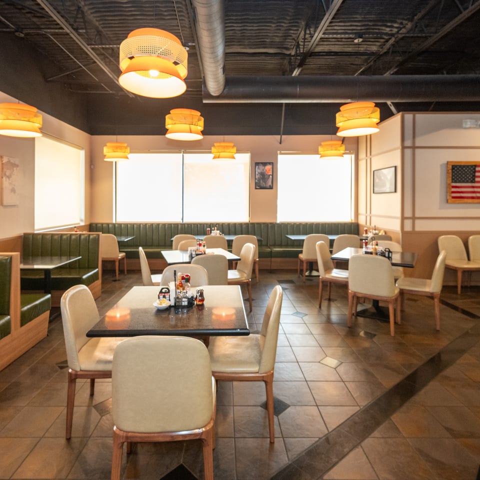 Spacious restaurant dining room with green booths, cream chairs, tiled floors, warm pendant lights, and an American flag on the wall.