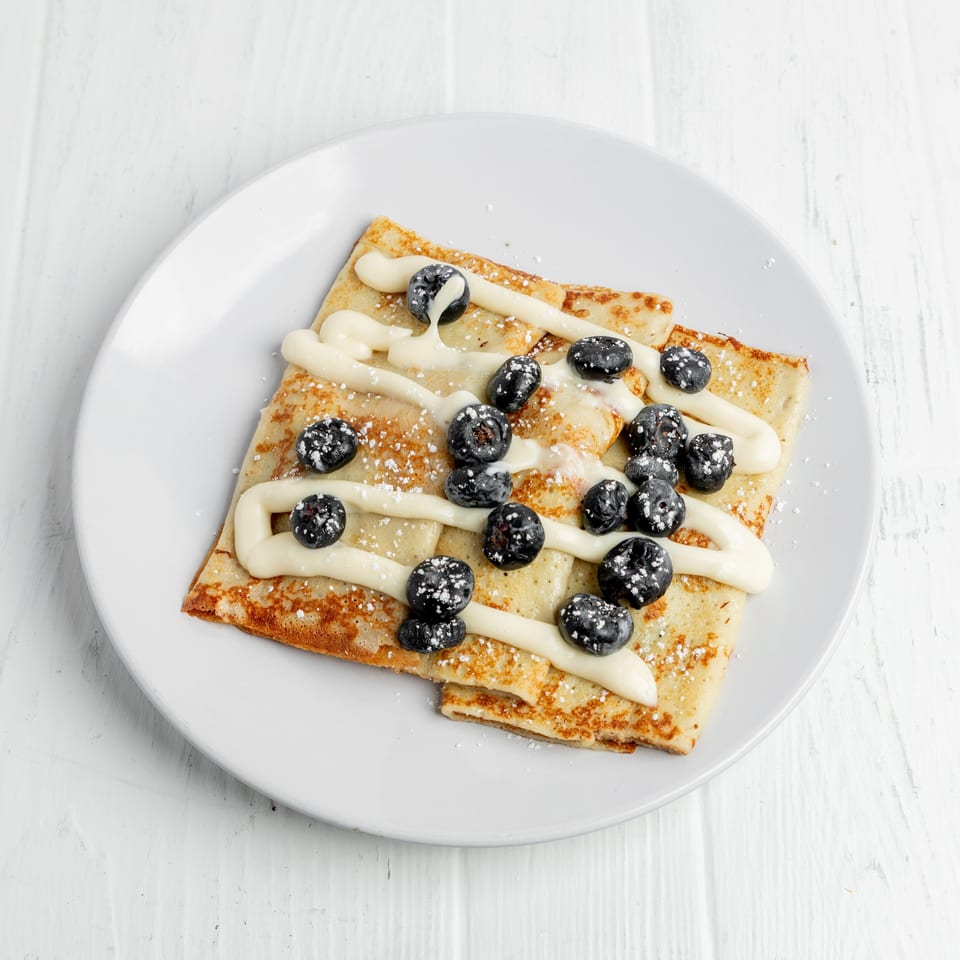Golden crepes topped with blueberries, vanilla cream, and powdered sugar on a white plate.
